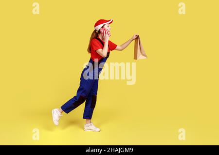 Side view portrait of delivery woman bringing door-to-door paper parcel to client and talking on phone with client, wearing overalls and red cap. Indoor studio shot isolated on yellow background. Stock Photo
