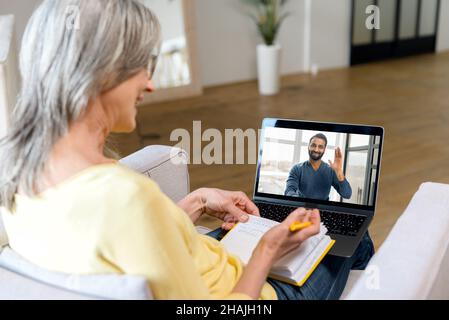 Smiling senior woman talking online with young Indian man on the laptop screen. Mature lady using laptop for video meeting with online tutor, taking notes, video call from home Stock Photo