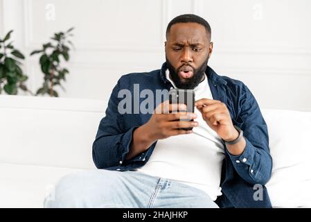 Shocked young African-American bearded guy in casual staring at smartphone screen sitting on the sofa at modern apartment, disappointed black guy received unexpected bad new Stock Photo