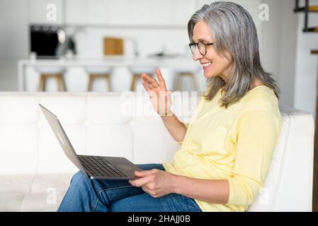 Pleased senior woman has video call on the laptop sitting on the couch at home, chatting with friends or family staying at home. Female teacher connecting with students, waving hello, greeting Stock Photo