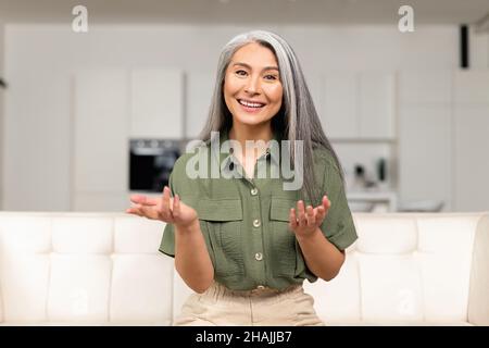 Webcam view at gray-haired woman holding video call sitting on the sofa at home, middle-aged Asian lady looks at camera and talking, involved virtual meeting, video screen Stock Photo
