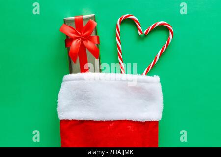 Christmas stocking with small gift box and candy canes on green background. Flat lay composition of holiday decorations. Stock Photo