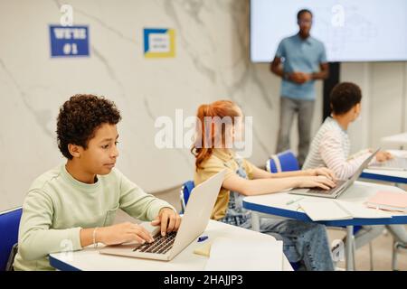 Side view portrait of teenge boy using laptop while studying in modern classroom with group of children, copy space Stock Photo
