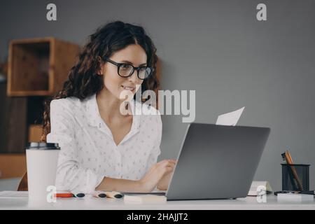 Happy young european woman wearing glasses in white shirt working on modern laptop from home Stock Photo