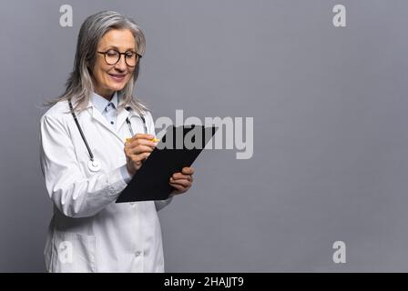 Senior doctor in white medical gown with stethoscope on shoulders taking notes standing with clipboard isolated on gray, mature physician therapist writing down treatment plan, writes a prescription Stock Photo