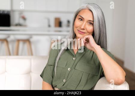 Close-up portrait of charming middle-aged woman with long silver hair sitting in relaxed athmosphere, mature 50s asian female resting at home, looking at camera and smiling with calm smile Stock Photo