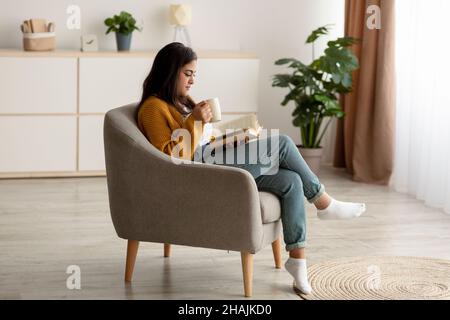 Attractive arab woman with cup of aromatic coffee and open book relaxing in armchair at home, having lazy weekend Stock Photo