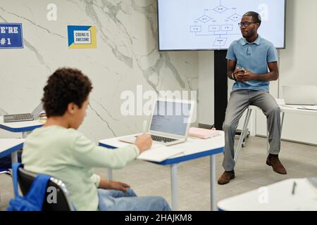 Full length portrait of young African-American teacher sitting on desk while teaching coding class for children, copy space Stock Photo