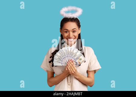 Portrait of satisfied smiling angelic woman with dreadlocks and nimb over head holding fan of dollars banknotes, wearing white shirt. Indoor studio shot isolated on blue background. Stock Photo