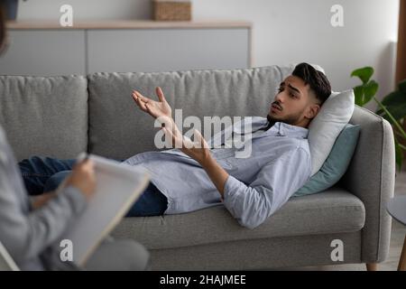 Depressed arab man lying on couch at psychologist's office, having session with counselor, seeking professional help Stock Photo