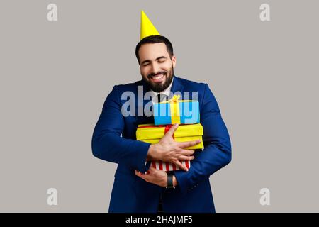 Extremely happy bearded man posing in party cone, holding embracing stack of presents, happy birthday, wearing official style suit. Indoor studio shot isolated on gray background. Stock Photo