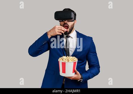 Portrait of excited amazed bearded businessman in VR headset watching movie with popcorn, eating tasty snack, wearing official style suit. Indoor studio shot isolated on gray background. Stock Photo