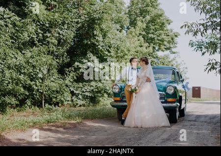 Just married wedding couple is standing near the retro vintage car in the park. Summer sunny day in forest. bride in elegant white dress with bouquet Stock Photo