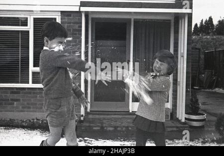 1960s, historical, a boy and his little sister throwing snowballs at each other outside the front of their home, a semi-detached suburban house, England, UK. Although it is wintertime, the boy is in a pair of shorts! Stock Photo