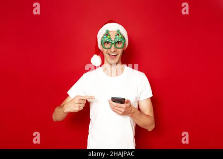 an excited man in fancy green glasses red hat points with his index finger at smartphone in his hand Stock Photo