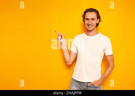 young curly haired smiling man in casual clothes points with his index finger at an empty area for advertising, promotions, discounts. isolated on yellow background with space for text. concept people. High quality photo Stock Photo