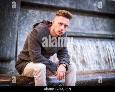 Young man sitting by fountain, looking at camera in city setting Stock Photo