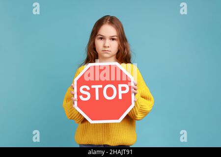 Little girl holding Stop symbol, showing red traffic sign, warning you about road safety rules, wearing yellow casual style sweater. Indoor studio shot isolated on blue background. Stock Photo