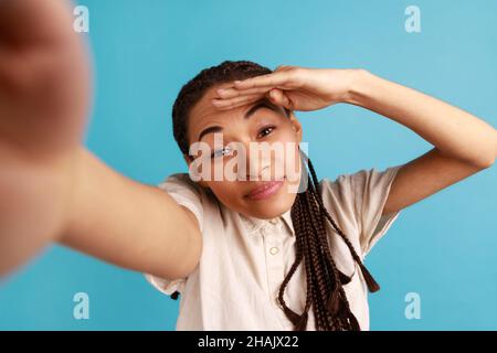 Beautiful woman with black dreadlocks has positive face expression, making selfie, looking far away, point of view photo, wearing white shirt. Indoor studio shot isolated on blue background. Stock Photo