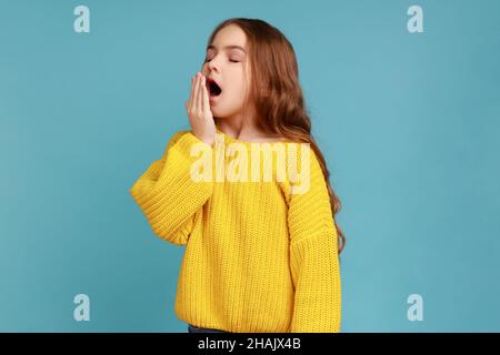 Little sleepy girl covering mouth with hand while yawning with eyes closed, drowsy child waking up, wearing yellow casual style sweater. Indoor studio shot isolated on blue background. Stock Photo