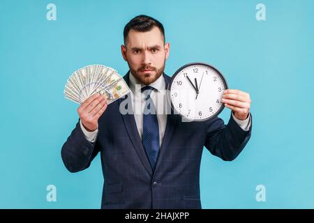 Time to make money. Sad man with beard wearing official style suit holding big clock and dollars banknotes, looking at camera serious facial expression. Indoor studio shot isolated on blue background. Stock Photo