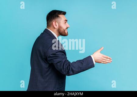 Hi, nice to meet you. Side view of friendly smiling handsome man wearing official style suit outstretching hand, offering handshake on job interview. Indoor studio shot isolated on blue background. Stock Photo