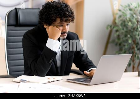 Frustrated, tired, overworked young Indian businessman, manager or IT professional with a beard sitting at the table in the office, frustrated by the news or financial loss, experiencing stress Stock Photo