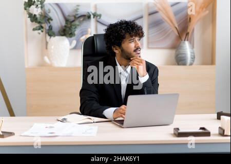 Pensive young successful curly-haired Indian ceo, top manager or businessman in formal suit sitting at table in modern office, look away, thinking over strategy, brainstorming, dreaming about vacation Stock Photo