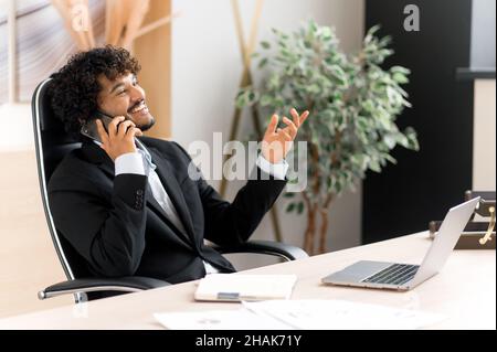 Successful joyful Indian businessman, marketer or manager, sitting at table in office, wearing stylish suit, talking with pleasure on his cell phone with his colleagues, smiling friendly, looking away Stock Photo