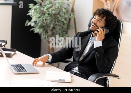 Successful happy Indian IT professional, marketer or manager, sitting at his desk in a formal suit, talking with pleasure on a smartphone while working, smiling friendly, looking away Stock Photo