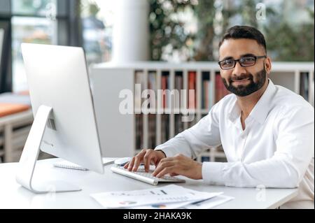 Smart successful handsome Indian businessman, manager, office worker with beard and glasses. Business owner sitting at table in modern office, using computer, looking at camera, smiling Stock Photo