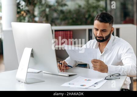Busy successful Indian business leader, manager or office worker with beard, sitting in an office, using computer, studying documents and diagrams, conducting analytics, taking notes Stock Photo