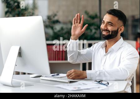 Happy friendly, confident successful Indian businessman or CEO with a beard, sitting in an office, wearing a white shirt, using a computer for personal communication, waving his hand, smiling Stock Photo