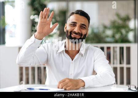 Headshot of confident joyful young adult Indian businessman, manager with beard, in white shirt, sit at a table in office, communicating colleagues via video communication, waving his hand, smiling Stock Photo