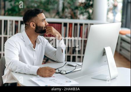 Smart self-confident successful Indian businessman, top manager or ceo with beard sits in an office, wears a white shirt, looks thoughtfully aside, ponders business strategy or project Stock Photo