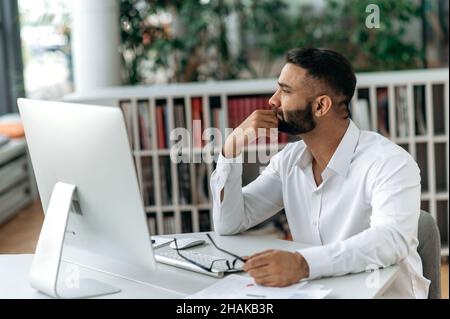 Pensive, intelligent, self-confident successful Indian businessman, top manager or ceo with beard sits at table, wears stylish shirt, looks to side, ponders business strategy or project. Stock Photo