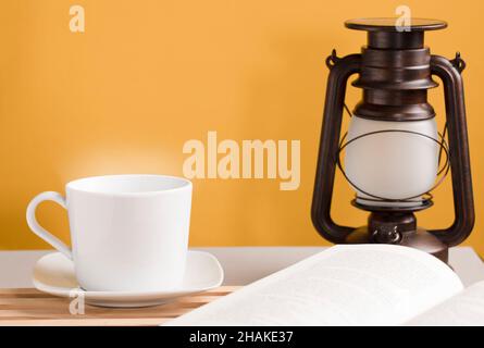 White cup of coffee with hot steam, on the desk with open book and sunlight lamp, orange-yellow background Stock Photo