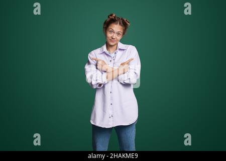 Woman holding hands crossed pointing index fingers aside, showing two directions wwith copy sapce. Isolated on green background. Gesturing Stock Photo