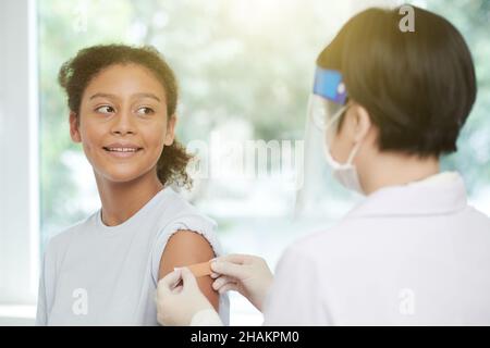 Smiling teenage girl looking at doctor sticking adhesive plaster on her arm after injecting vaccine Stock Photo