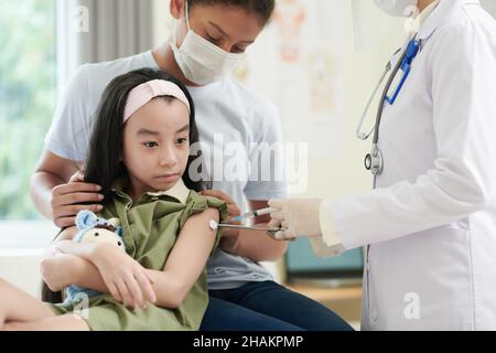 Elder sister reassuring little girl getting shot of vaccine against coronavirus in clinic Stock Photo
