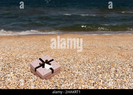 A box with a gift tied with a ribbon lies on the beach near the sea. Front view, vertical. Stock Photo