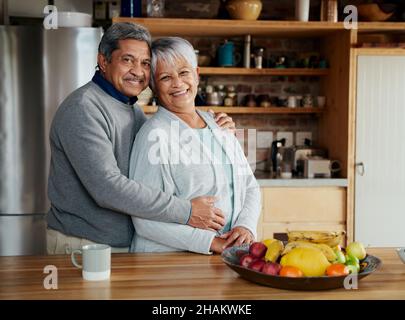 Portrait of happily retired elderly biracial couple standing, hugging and smiling at camera while in kitchen. Stock Photo