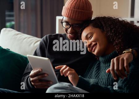 Cute adult couple, multi-ethnic, smiling at tablet on sofa in modern apartment Stock Photo