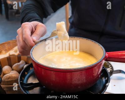 A man’s hand holds on a fork a piece of bread that has been dipped in a cheese fondue pot Stock Photo