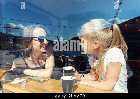 Through window view of young adult happy mom enjoy having fun talking, drink hot chocolate laugh with cute little daughter sitting at cafe restaurant Stock Photo
