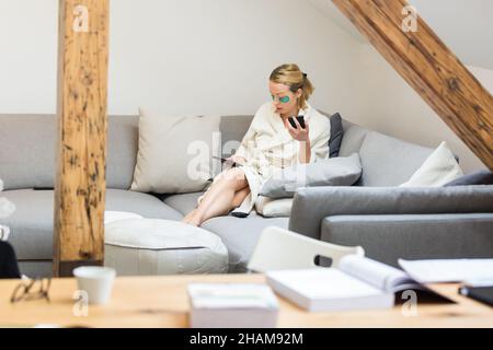 Young female entrepreneur wearing beauty face mask and cosy warm bathrobe in the morning, working remotly from her living room sofa. Remote work from Stock Photo