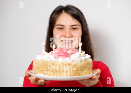 A brunette woman in red clothes holds a large white cake with candles, makes a wish Stock Photo