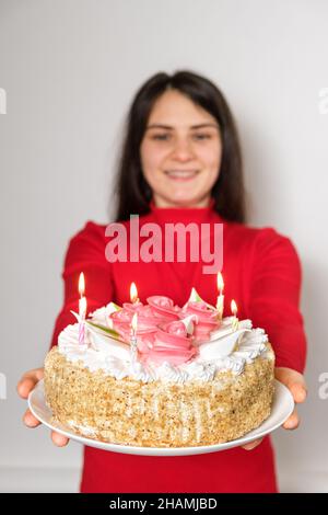 A brunette woman in red clothes holds a large white cake with candles, makes a wish Stock Photo