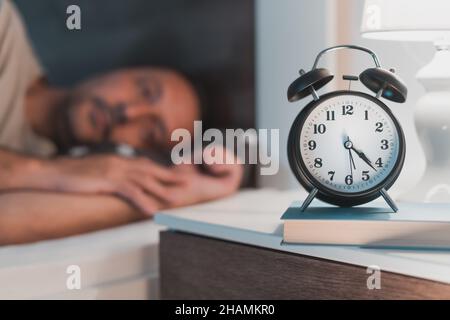 Sleepless man lying in bed and looking at alarm clock on bedside nightstand, selective focus Stock Photo