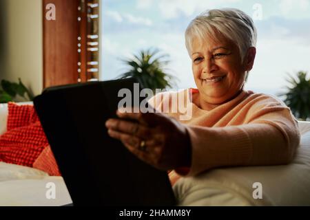Multi-cultural elderly female smiling while using electronic tablet, sitting on living room sofa in the daytime. Stock Photo